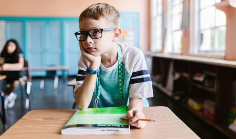 A young boy wearing eyeglasses sits at a desk in a classroom, focused on a book.