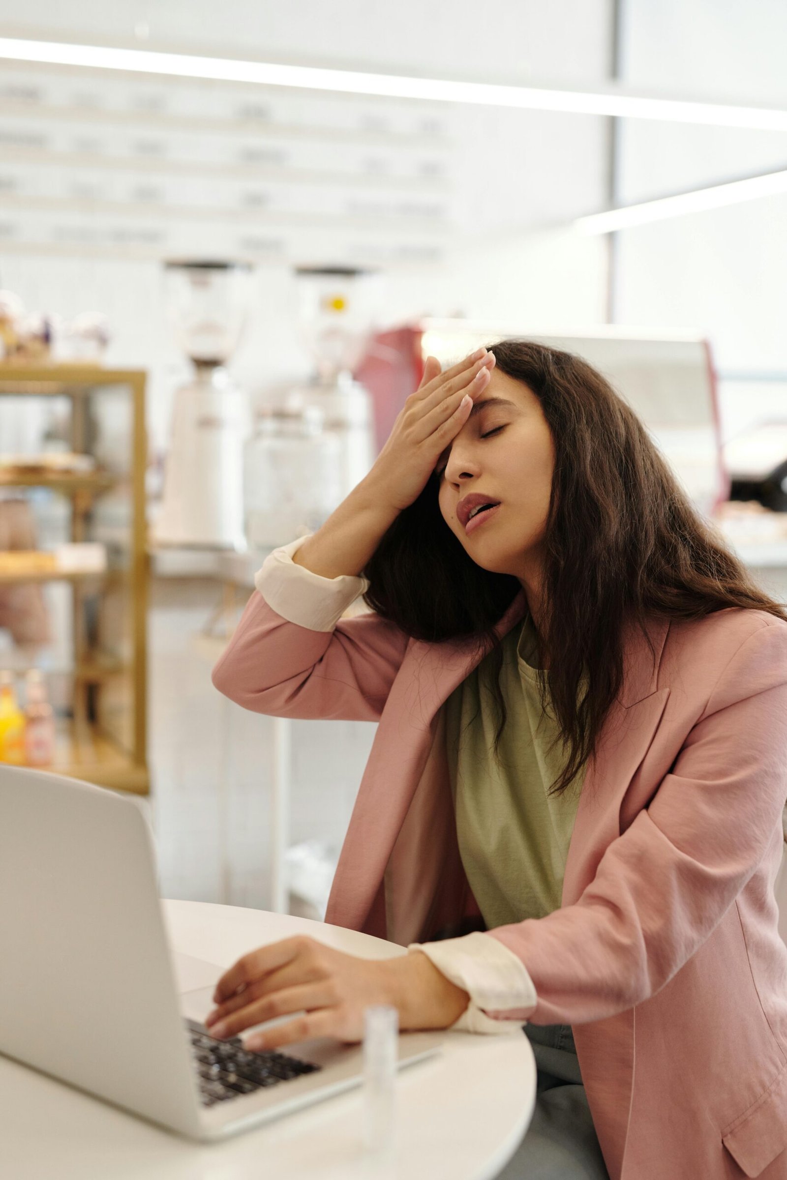 A young woman in a pink blazer looks unwell while working on a laptop in a cafe.